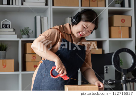 Woman in denim apron using scissors to cut packing tape in a modern workspace, preparing boxes for shipment Woman in denim apron using scissors to cut packing tape in a modern workspace, preparing boxes for shipment 122716837