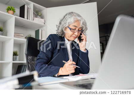 Elderly business woman managing tasks while on the phone in a modern office setting Elderly business woman managing tasks while on the phone in a modern office setting 122716978