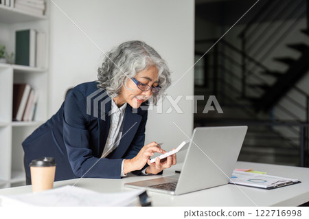 Elderly businesswoman calculating figures while engaged with a laptop in her modern office 122716998