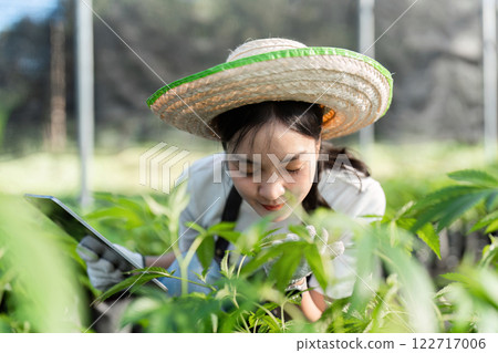 Young Female Farmer Examining Plants in a Greenhouse with a Tablet and Straw Hat 122717006
