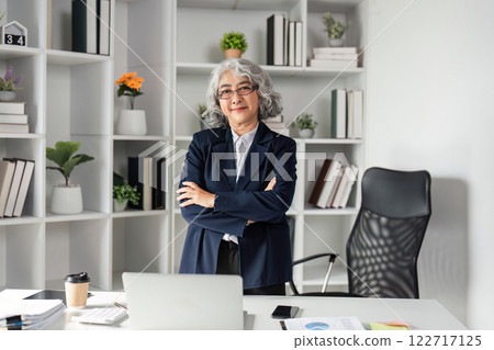 Senior female business executive confidently standing with arms crossed in a stylish office environment Senior female business executive confidently standing with arms crossed in a stylish office environment 122717125
