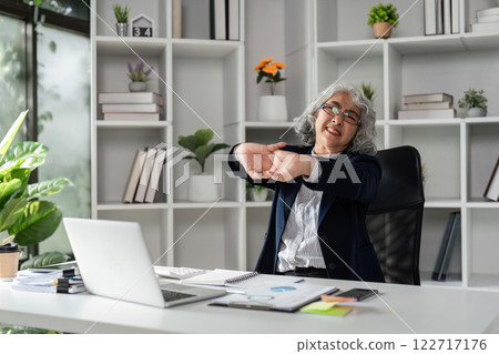 Cheerful elderly businesswoman stretching at her desk, enjoying a moment of relaxation in a vibrant office 122717176