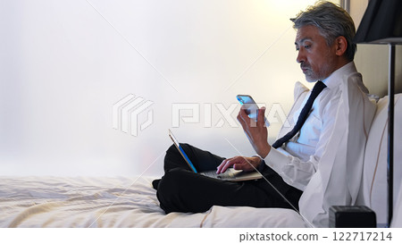 Male business person looking at smartphone and laptop in bed 122717214