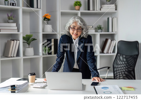 Confident Elderly Businesswoman in Modern Office Setting with Laptop and Documents Confident Elderly Businesswoman in Modern Office Setting with Laptop and Documents 122717455