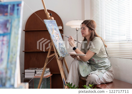 Young Asian female artist is intently painting acrylic paints on canvas in a painting studio. 122717711