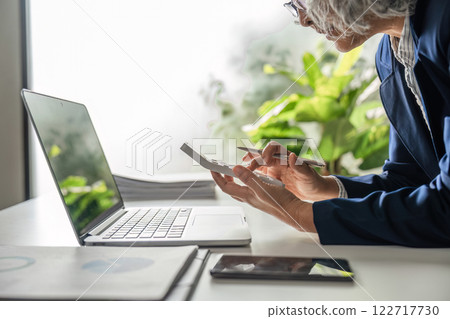 Elderly businesswoman reviewing financial data on a calculator and laptop in her stylish office 122717730