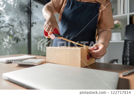 Woman in denim apron taping a cardboard box in a bright home office, preparing it for dispatch Woman in denim apron taping a cardboard box in a bright home office, preparing it for dispatch 122717814