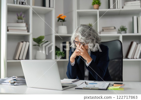 Tired elderly businesswoman with hands on her face while working at her desk in a contemporary office Tired elderly businesswoman with hands on her face while working at her desk in a contemporary office 122717863