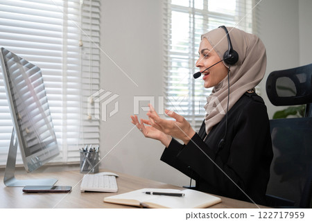 Call center worker, young Muslim woman wearing hijab, talking to customer on call phone on computer in customer service office Call center worker, young Muslim woman wearing hijab, talking to customer on call phone on computer in customer service office 122717959