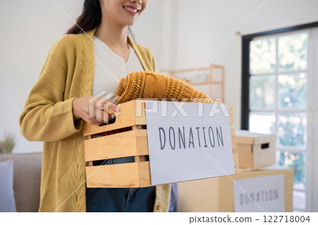 A cute Asian woman stands holding a box, sorting unwanted clothes for donation. 122718004
