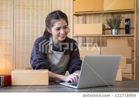 Young Asian female online business entrepreneur is searching customer information on laptop and packing products on table to ship to customer. Young Asian female online business entrepreneur is searching customer information on laptop and packing products on table to ship to customer. 122718009
