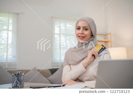 A young Muslim woman wearing a hijab sits contentedly shopping on her laptop, paying through an online banking app and holding a credit card. 122718123