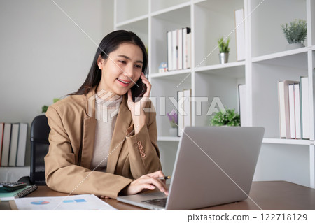 Young Asian business woman sits on the phone in an online business meeting using a laptop in a modern home office decorated with shady green plants. 122718129