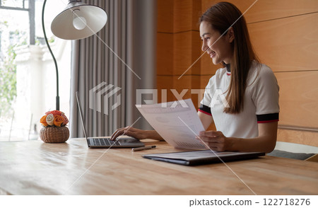 Young Woman Focused on Paperwork in a Modern Room with Laptop and Natural Light 122718276