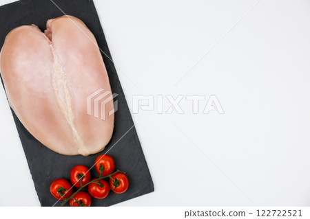 Close-up view from top left: chicken fillet with a sprig of tomatoes next to it lies on a black stone board on a white background with space for text. The concept of cooking, dietary menu, healthy and 122722521