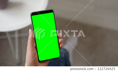 Young woman sitting at home holding smartphone green mock-up screen in hand. Female person use chroma key mobile phone. Vertical mode. Touching, swiping display, tapping, surfing internet social media Young woman sitting at home holding smartphone green mock-up screen in hand. Female person use chroma key mobile phone. Vertical mode. Touching, swiping display, tapping, surfing internet social media 122726425