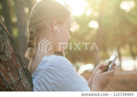 Happy Teenage girl in white shirt and headphones typing on mobile phone and standing Happy Teenage girl in white shirt and headphones typing on mobile phone and standing 122726985