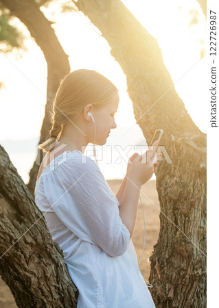 Beautiful teenage girl in white skirt in headphones holding mobile phone and standing sideways Beautiful teenage girl in white skirt in headphones holding mobile phone and standing sideways 122726987