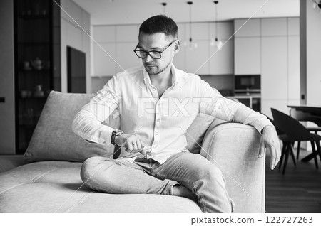 Busy man checking time on his watch. Young man waiting for important meeting. Handsome male in casual clothes sitting on a couch. Black and white image. 122727263