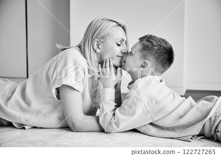 Little son showing love to his mom. Young mother and her kid lying on white carpet and spending time together at home. Lovely afternoon with family. Black and white image. 122727265
