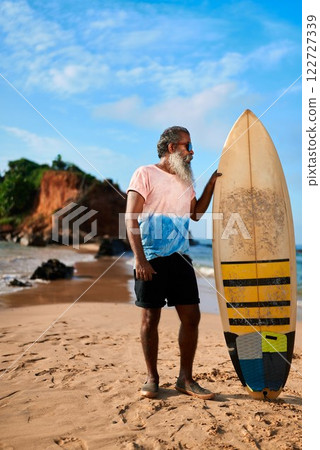 Senior man with surfboard stands on sandy beach. Elderly surfer poses under blue sky by rocky coast. Active retiree enjoys outdoor sea adventure. Tropical surf lifestyle travel concept. 122727339