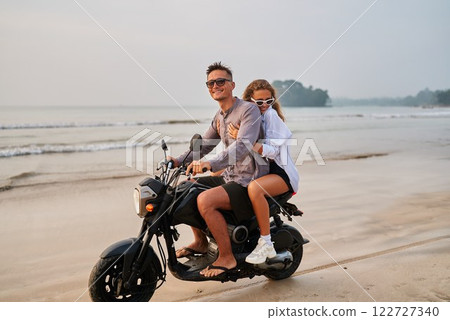 Couple rides motorbike along beach. Man drives scooter, woman sits behind, enjoying sunny ocean view. Travel adventure, exploring seaside, sand, tropical vibe. Perfect for travel tours, holiday ideas. 122727340