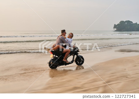 Couple on motorbike riding along sandy beach at sunrise. Romantic escape, joyful exploration. Man, woman in love, sharing adventure. Motorcycle, ocean, holiday vibe, freedom. 122727341