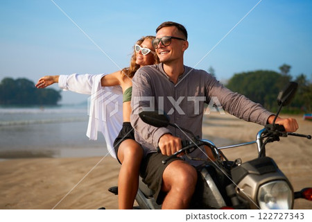 Happy couple rides motorbike on scenic coastal beach. Man drives scooter. Woman spreads arms enjoying wind and sun. Tropical holiday vibe. Perfect for travel, adventure, freedom themes. Happy couple rides motorbike on scenic coastal beach. Man drives scooter. Woman spreads arms enjoying wind and sun. Tropical holiday vibe. Perfect for travel, adventure, freedom themes. 122727343