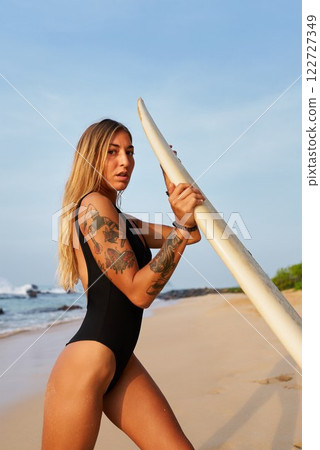 Young woman with tattoos holding surfboard on sandy beach. Female surfer prepares for surfing lesson by ocean. Empowerment, active lifestyle, outdoor adventure for women in water sports. Young woman with tattoos holding surfboard on sandy beach. Female surfer prepares for surfing lesson by ocean. Empowerment, active lifestyle, outdoor adventure for women in water sports. 122727349