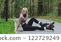 Cute blonde puts on roller skates. She feels free and light while roller skating alone in the city park. Stunning Caucasian woman with a beautiful figure uses roller skates for outdoor sports. 122729249