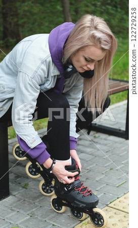 Cute blonde puts on roller skates. She feels free and light while roller skating alone in the city park. Stunning Caucasian woman with a beautiful figure uses roller skates for outdoor sports. 122729258