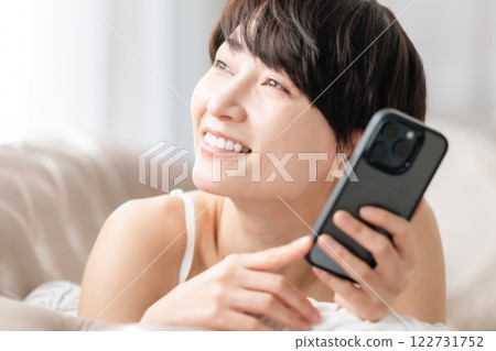 Smiling young woman holding a smartphone in her living room Smiling young woman holding a smartphone in her living room 122731752