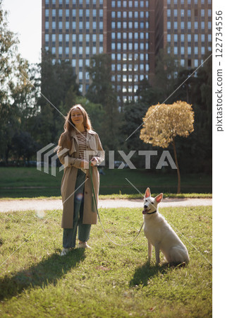 young woman walks with a white dog in park. lady in beige raincoat hugs husky on green lawn young woman walks with a white dog in park. lady in beige raincoat hugs husky on green lawn 122734556