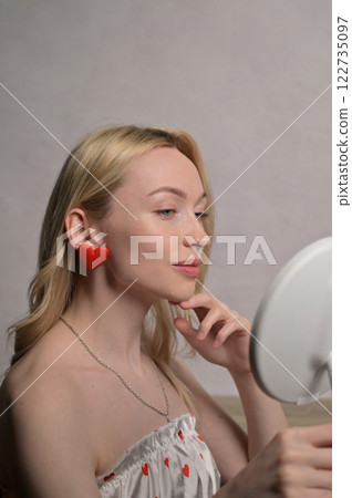 Blonde smiling girl in a white pajama heart pattern with red heart shape earring look at round mirror near wooden table. White wall background. Valentine's Day preparation Blonde smiling girl in a white pajama heart pattern with red heart shape earring look at round mirror near wooden table. White wall background. Valentine's Day preparation 122735097