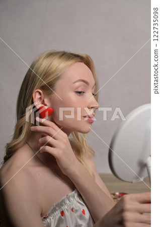 Blonde smiling girl in a white pajama heart pattern touching a red heart shape earring look at round mirror near wooden table. White wall background. Valentine's Day preparation Blonde smiling girl in a white pajama heart pattern touching a red heart shape earring look at round mirror near wooden table. White wall background. Valentine's Day preparation 122735098
