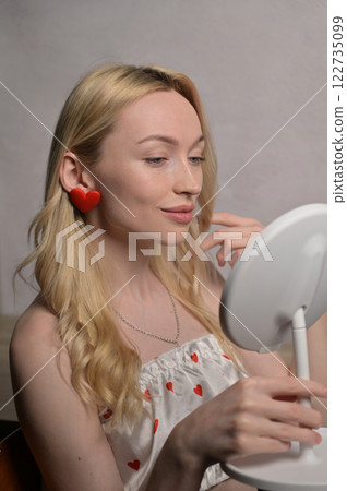 Blonde smiling girl in a white pajama heart pattern with red heart shape earring touching hair and look at round mirror near wooden table. White wall background. Valentine's Day preparation 122735099