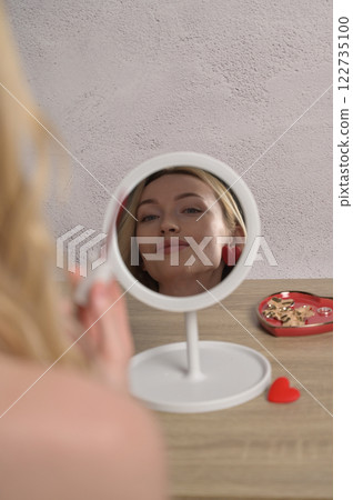 Reflection blonde smiling girl in the round mirror. Heart shape jewelry on the wooden table, white wall background. Makeup preparation for Valentine's Day 122735100