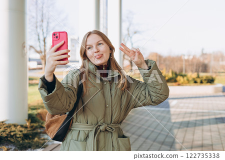 Laughing young woman taking selfie on the urban background. Cute beautiful woman using mobile phone. 30s girl recording self video, talking on online call 122735338