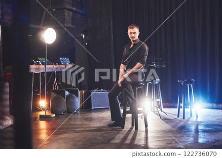 Magic look. Handsome young man in black clothes looking at camera while sitting on chair near the in dark room with light Magic look. Handsome young man in black clothes looking at camera while sitting on chair near the in dark room with light 122736070