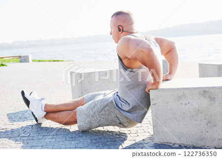 Profile view of a strong man exercising his arms and doing tricep dips outdoors in a park bench Profile view of a strong man exercising his arms and doing tricep dips outdoors in a park bench 122736350