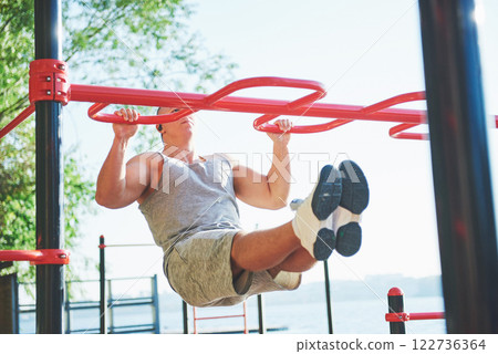 Muscular man with beautiful torso exercising on horizontal bars on a blurred park background. Young man doing pull-ups outdoors 122736364