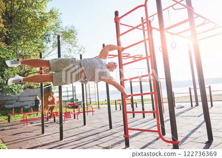 Muscular man with beautiful torso exercising on horizontal bars on a blurred park background. Young man doing pull-ups outdoors 122736369