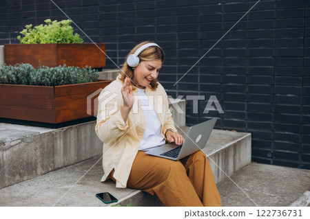 Happy 30s woman in headphones doing business on laptop and smiles. Young woman enjoy her work outdoor. Remote work, freelance. Young pretty female talking on video call and waving hand on stairs Happy 30s woman in headphones doing business on laptop and smiles. Young woman enjoy her work outdoor. Remote work, freelance. Young pretty female talking on video call and waving hand on stairs 122736731
