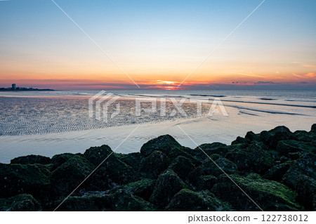 A tranquil sunset unfolds beautifully over calm waters and a picturesque rocky shoreline 122738012