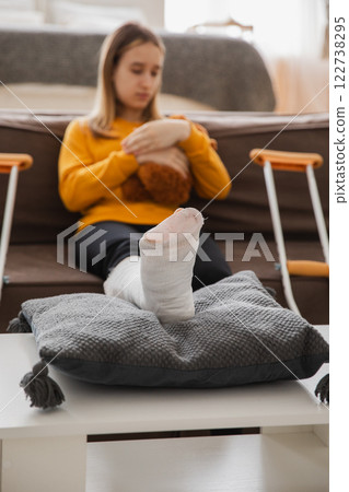 Broken leg. Young girl with broken leg cast sitting on sofa. Sad little kid recovering from broken leg at home. Girl using cast and crutches for rehabilitation. Accident injury fracture treatment 122738295