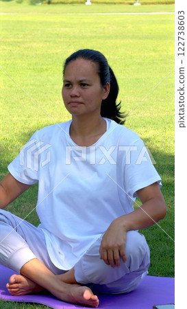 Asian woman practicing yoga outdoors, surrounded by serene green hues, radiating calm, inner peace, symbolizing tranquil lifestyle focused on wellness. 122738603