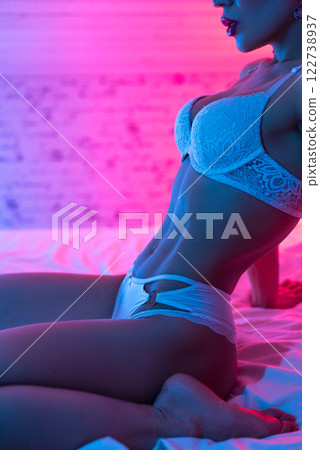 A woman with a sexy body in beautiful white lingerie poses erotically on a bed in neon light A woman with a sexy body in beautiful white lingerie poses erotically on a bed in neon light 122738937