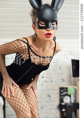 Sexy girl in a short black dress and a rabbit mask from a sex shop poses erotically in a mesh bodysuit 122738998