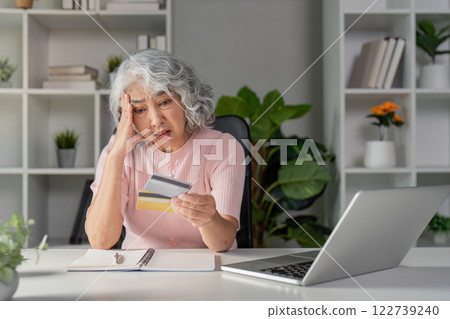 Concerned senior woman evaluating her credit cards while feeling stressed about online shopping in her home office Concerned senior woman evaluating her credit cards while feeling stressed about online shopping in her home office 122739240