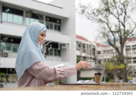 Muslim businesswoman in hijab smiling while working on a laptop outdoors with a coffee. Muslim businesswoman in hijab smiling while working on a laptop outdoors with a coffee. 122739365